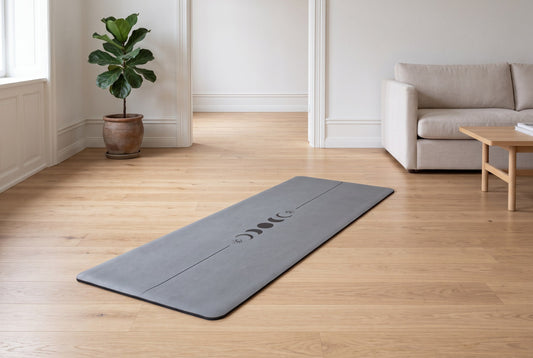 Graue Yogamatte, ausgerollt auf Holzboden in einem hellen Wohnraum mit Sofa und Zimmerpflanze