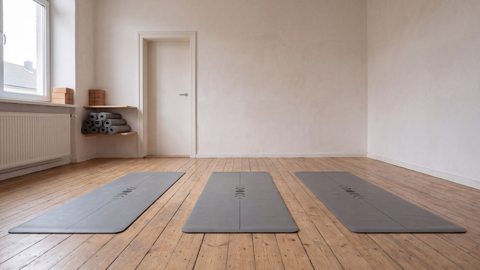Leerer Kursraum mit drei ausgerollten Movasana-Yogamatten auf Holzdielen, Regal mit gerollten Matten und Kork-Blöcken
